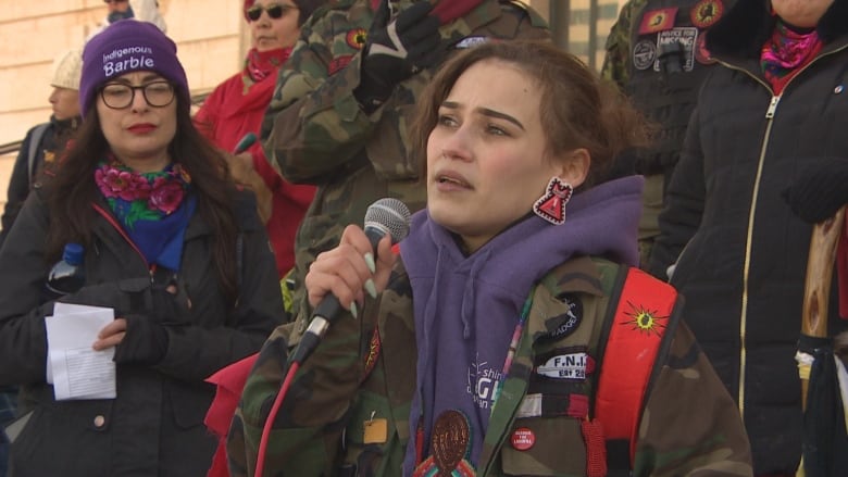 A young woman wearing a camouflage jacket and a red backpack speaks into a microphone.