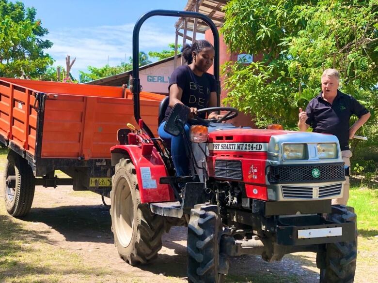 A woman drives farm equipment while a man in a dark shirt stands nearby, supervising