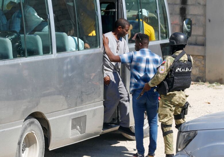 A man in a short-sleeved plaid shirt and jeans leans against the door of a silver bus as a second man wearing a grey sweat suit steps off the bus while a man in a military uniform holding a gun looks on. 