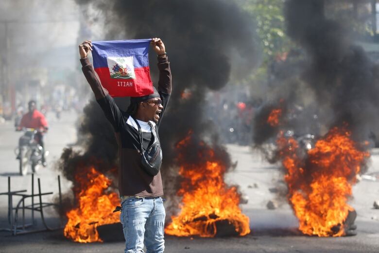 A man yells in the street while holding a Haitian flag above his head with both hands. Behind him, three piles of tires burn and release billowing columns of black smoke.