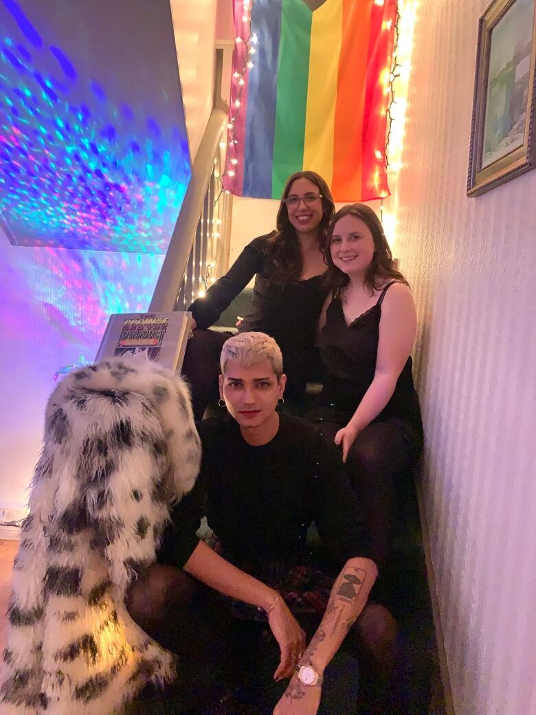 Godara with two other friends in a staircase, with a pride flag in the background.