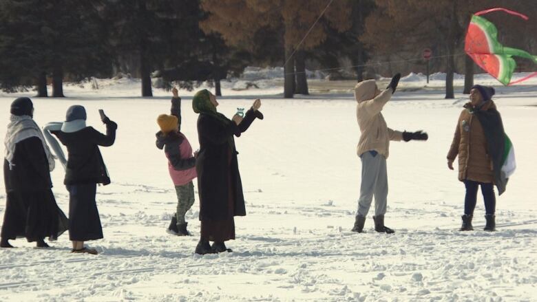 Half a dozen people stand in a snowy field, looking upward, with strings stretching upward. A low-flying kite is on the right of the image.