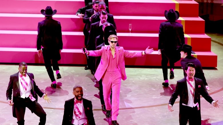 Ryan wears a pink suit with his arms stretched out, as other Kens surround him onstage.