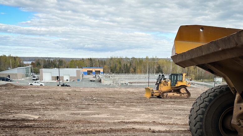 Yellow bulldozers scrape the earth in the foreground, while a new building and sewage treatment facility are built in the background.