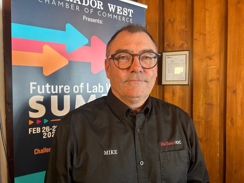 A man wearing a black dress shirt stands in a room with wood paneling on the walls and a banner for the Future of Lab West Summit.