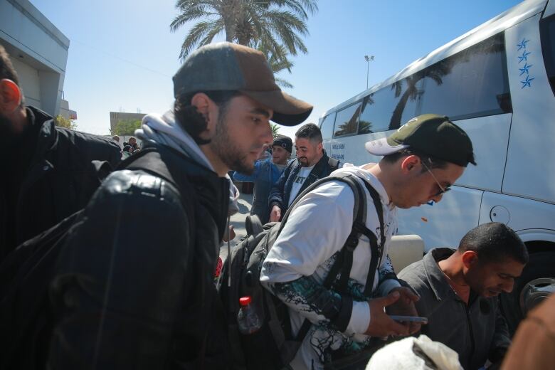 Two brothers wearing baseball caps and carrying backpacks wait to board a white tour bus.