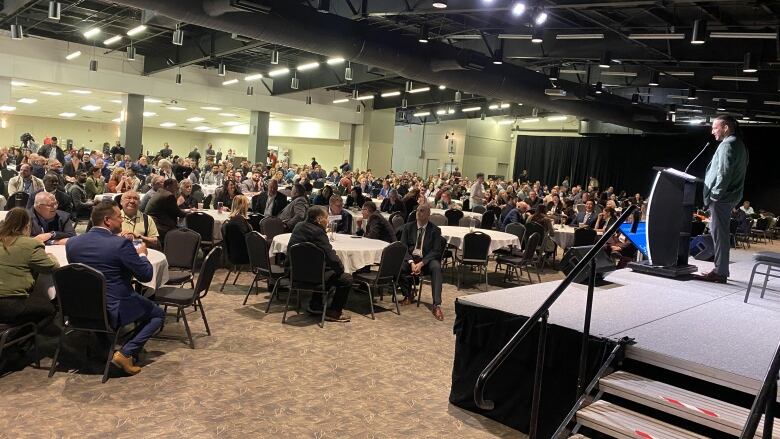 People sit at tables while a speaker talks on a stage.