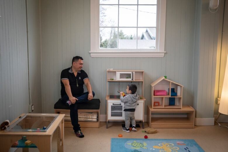 A toddler boy plays at a toy kitchen with his father sitting next to him looking fondly.