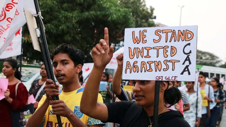 A group of young people stand outside holding signs. One in front, in hand-drawn block letters, reads: 