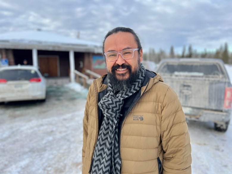 A smiling man with a beard and a scarf stands in a small parking lot in winter.