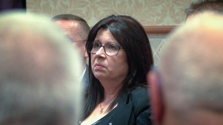 A middle-aged woman with a dark jacket and glasses sits among a crowd of people.