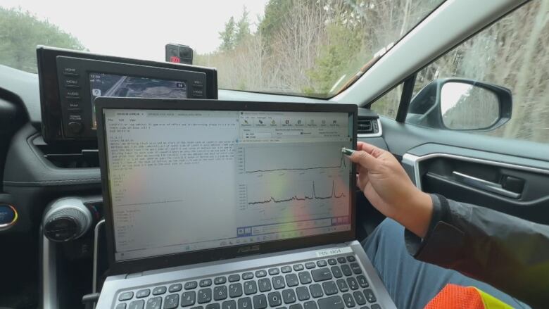 The screen of a laptop, viewed from inside a moving vehicle. A woman's hand is in the picture, pointing with her thumb at a blue line on the laptop screen. Underneath the woman's thumb is a spike in the line. 