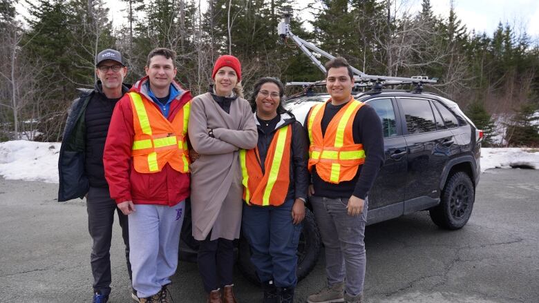 Five people stand in front of a black SUV with a metal antennae arm on its roof. There are three men and two women in the picture. Three of the people are wearing reflective orange safety vests. 