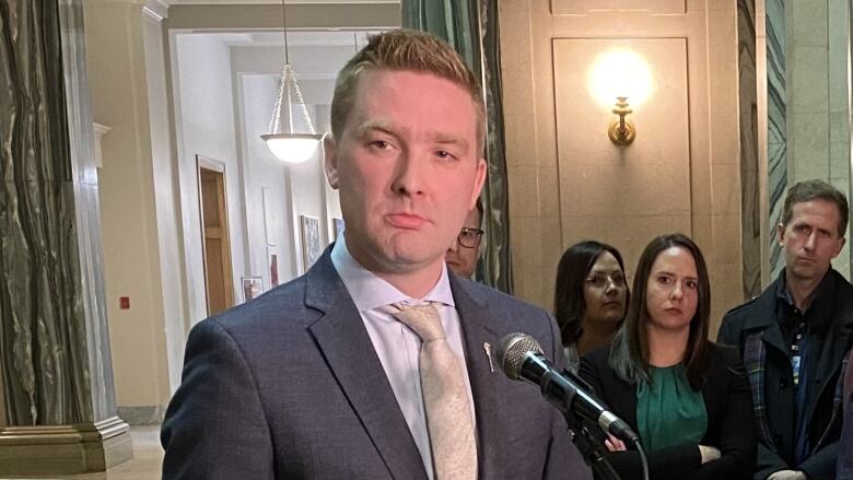 A man in a grey suit and a gold tie stands in front of a microphone. Over his right shoulder a woman in a green shirt and a black sweater crosses her arms. 