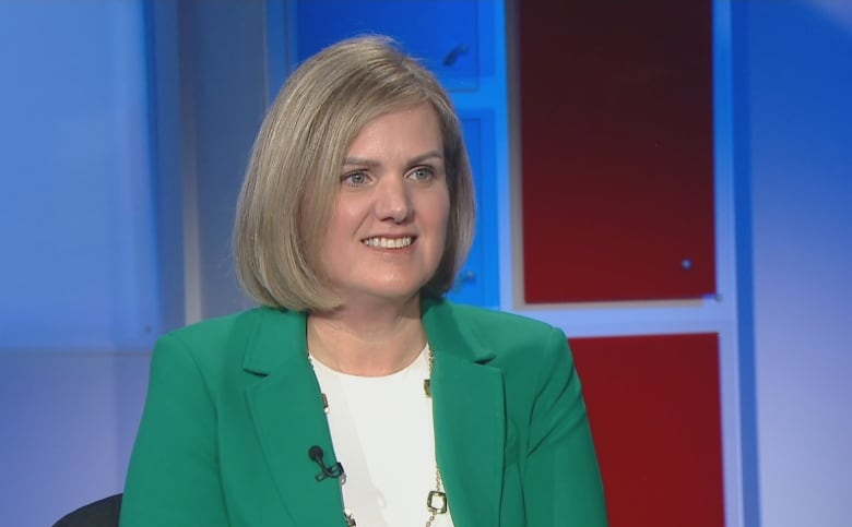 A woman in a green blazer and white shirt sits in a television studio.