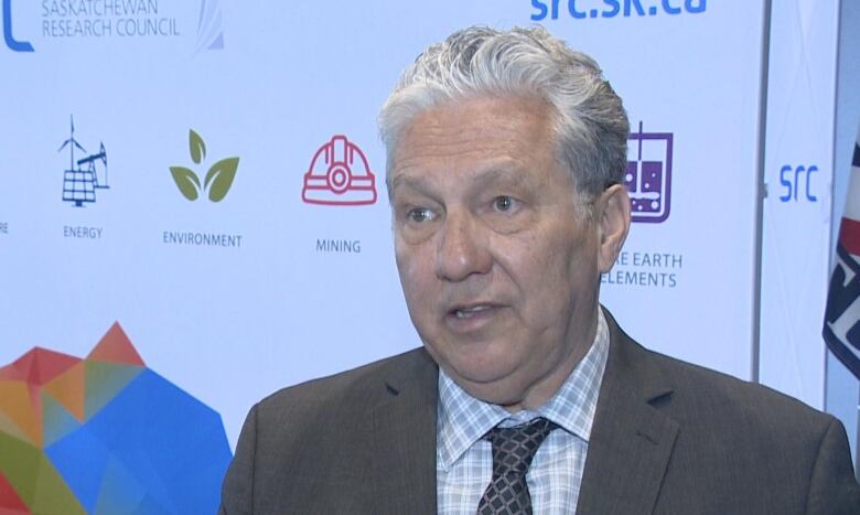 A man with grey-white hair, wearing a grey suit, is speaking.