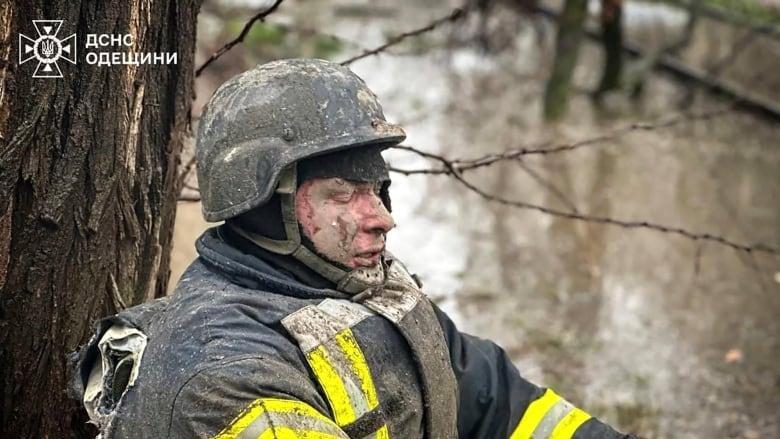 A man, his face caked in dirt, closes his eyes and leans against a tree. His uniform is tattered on its back.