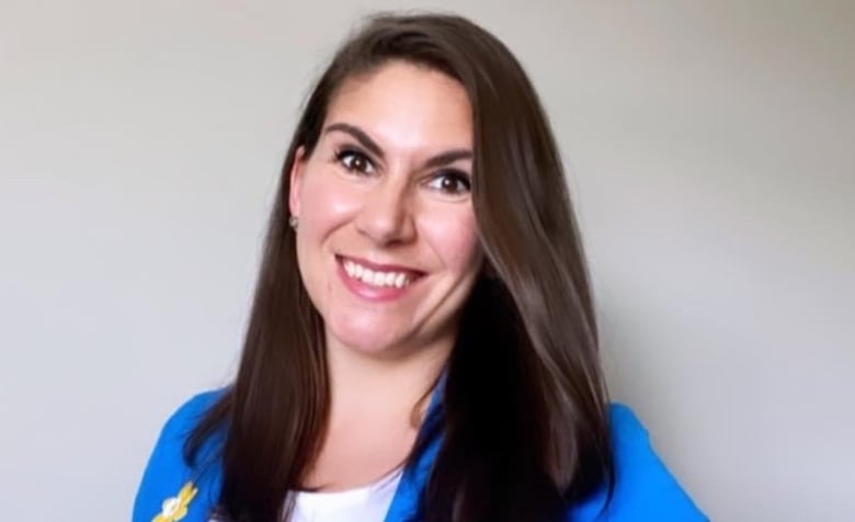 A woman in about her 30s with long brown hair and brown eyes smiles at the camera wearing a bright blue blazer with a yellow daffodil emblem on one lapel over a white t-shirt in an upper body portrait against a plain off-white background.