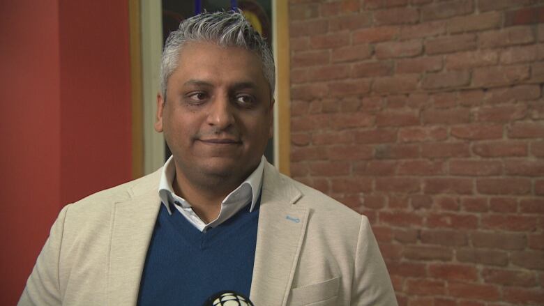 A man with grey hair wearing a tan blazer stands near a brick wall.