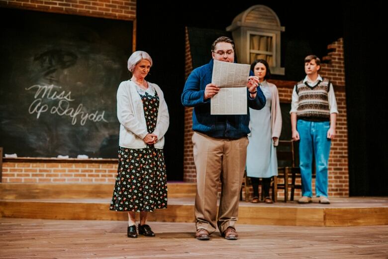 a woman on the left, a man standing in front of her reading a newspaper. 