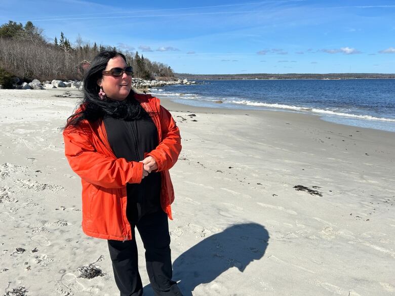 A woman with long, dark hair is wearing shades and a red-orange jacket. She's standing on a beach.