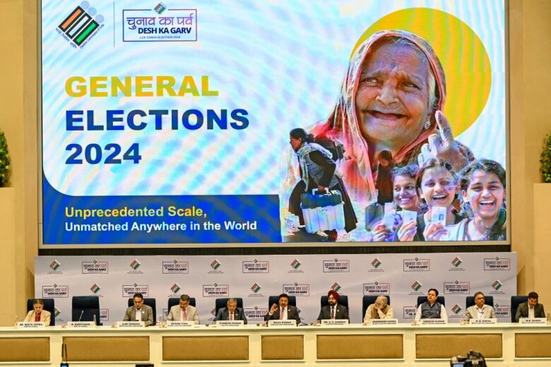 Members of India's election commission sit in a row in front of a sign announcing the country's national election.