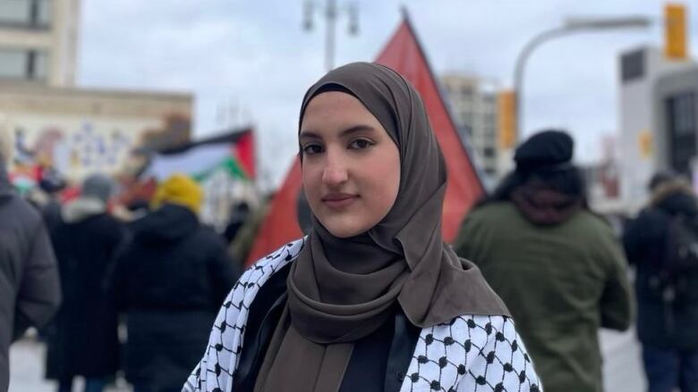 A woman standing in front of a crowd. A person behind her can be seen waving the Palestine flag.