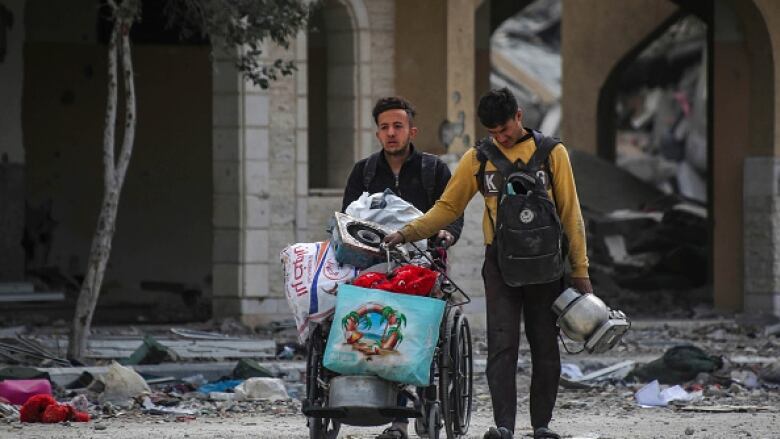 Two men walk amid rubble carrying belongings. The man on the left pushes a wheelchair filled with bags.