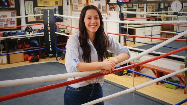 A smiling woman with long brown hair leaning on the inside of a boxing ring.