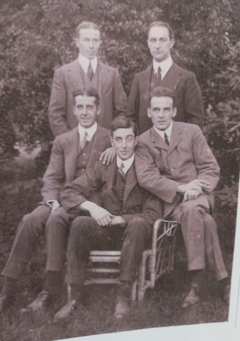 A group of five men sit and stand in front of some bushes. All men are dressed finely, in jackets and suits. The photo is more than 100 years old.