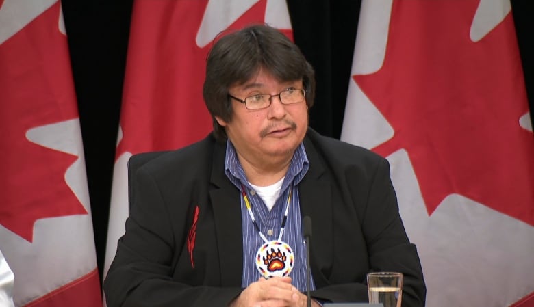 A man in a black suit sitting behind a desk with Canadian flags in the background. 