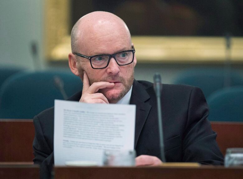 A bald white man holding a sheet of paper puts his hands on his cheek while sitting in front of a mic.