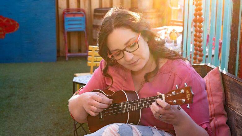 A woman with long brown hair and glasses sits in a chair and strums a ukelele.