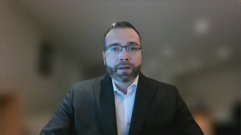 Man with dark hair and beard, wearing glasses and a black suit with white shirt.