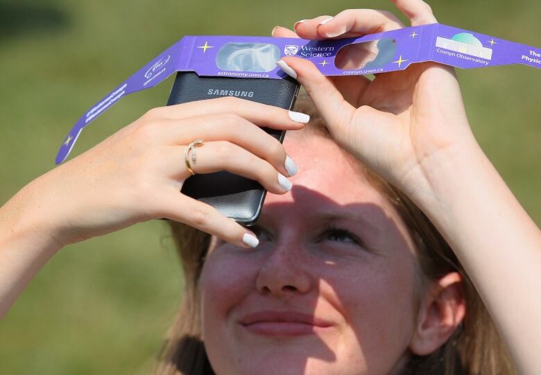 A girl holds solar eclipse glasses over the lens on her phone to take a picture of the sun in the sky during a partial eclipse.