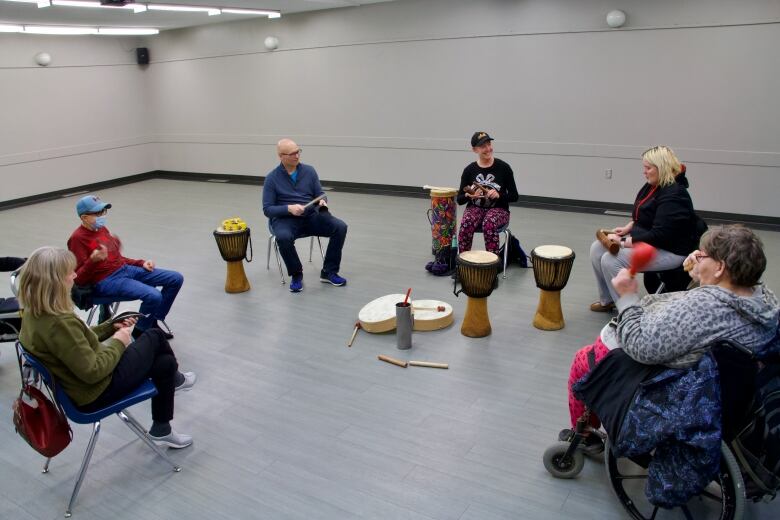 Six people sit in a circle. There is a pile of percussive instruments in the middle of them, and they are each holding one, such as wood blocks, a shaker and tambourine.