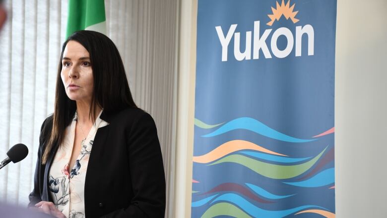 A woman with long dark hair wearing a black blazer and floral shirt underneath stands in front of a banner that says 