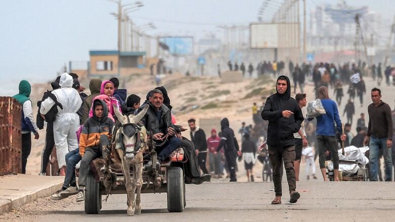 Dozens of people are shown walking along a dirt road. In the foreground, a small vehicle led by an animal is shown.