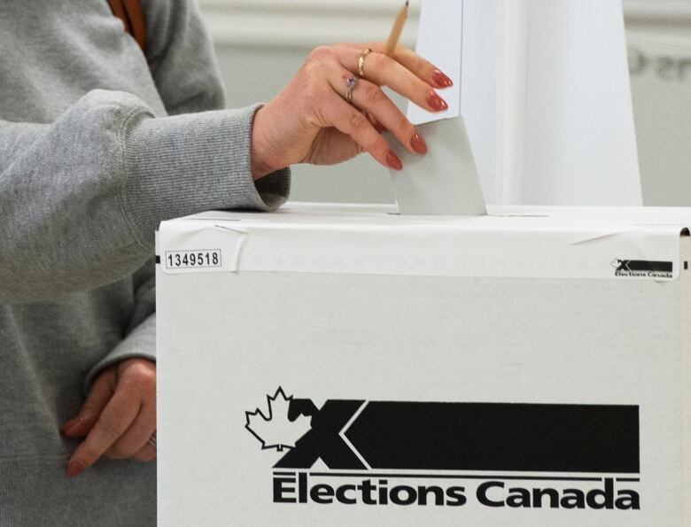 A hand holding a pencil puts a ballot into a voting box.