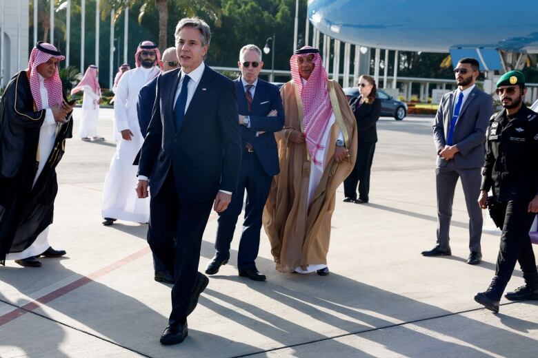 Men in suits and ties and other men in robes and headdresses are shown on the tarmac of an airport, with a portion of an airplane shown in the background.