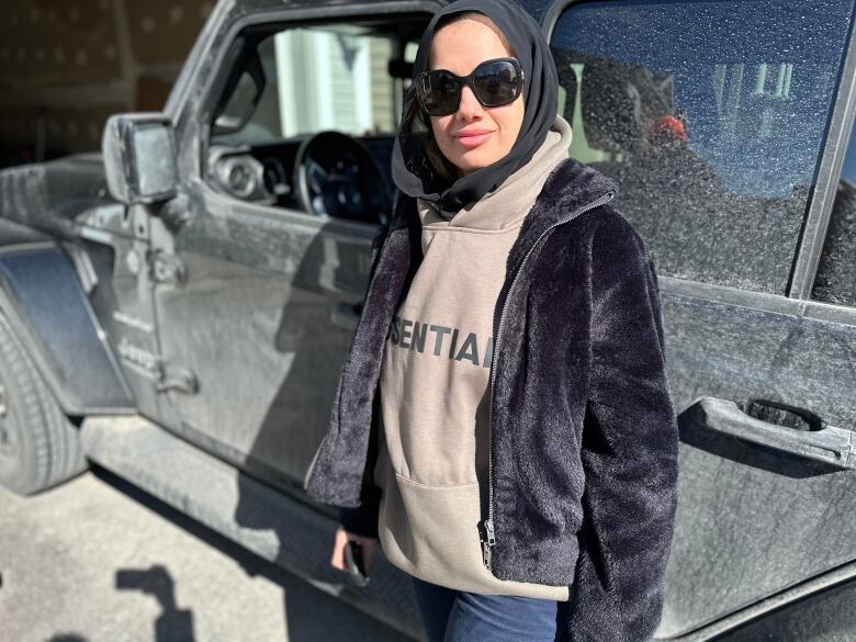 A woman with sunglasses stands in a black jacket in front of a Jeep.