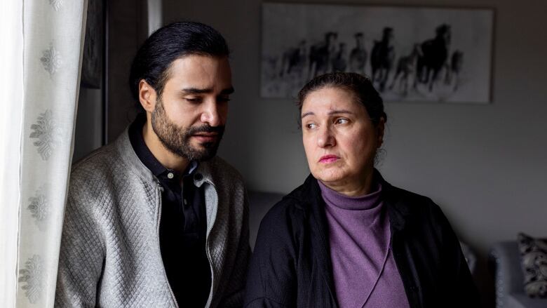 A man and an older woman stand-by-side next to a window. The man has his eyes cast downward and the woman looks to one side. Both are frowning, 