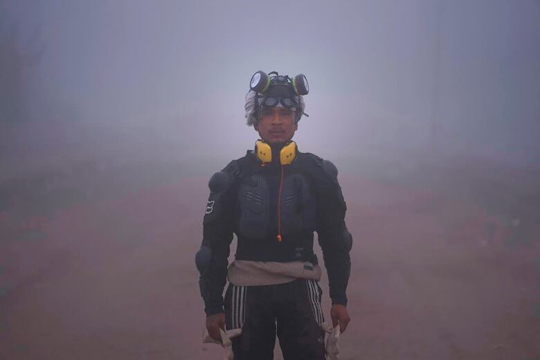 A man wearing a gas mask on his head, yellow ear protectors around his neck and padded or armoured clothing stands looking at the camera.