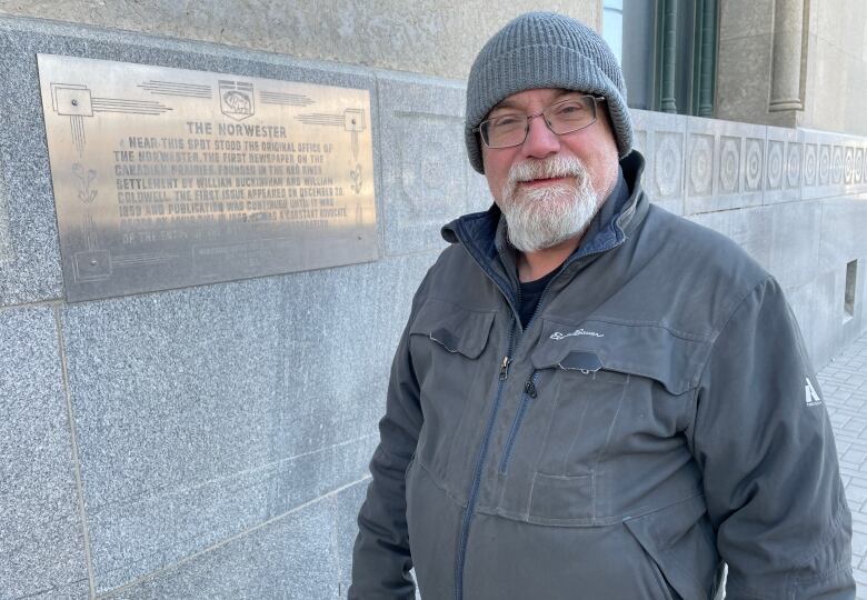 A man with a white goatee, and wearing a grey tuque, stands beside a memorial plaque mounted on a wall.