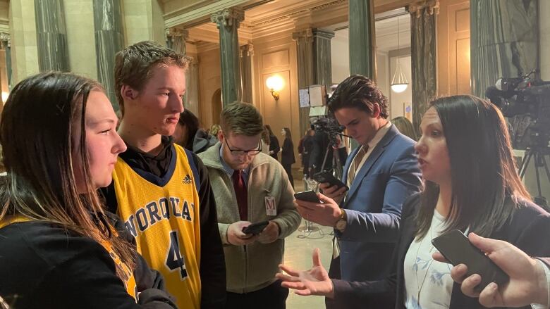 A woman in a suit, a boy in a yellow jersey and a girl also in a yellow jersey are standing inside the legislature. 