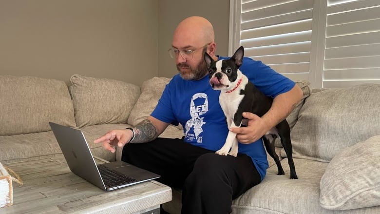 A man sits on a sectional sofa in front of a laptop with his dog under one arm.