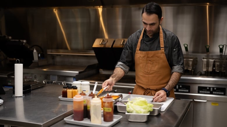 A man in a grey button-down and an orange chef's apron prepares to spread sauce on a hamburger bun inside a kitchen.