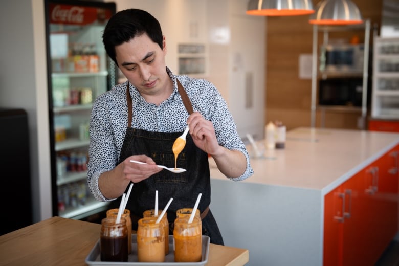 A man in a blue button-down shirt and a blue apron prepares to taste a spoonful of spicy sauce.