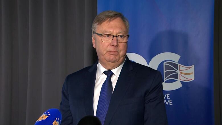 A man wearing a suit stands in front of a banner for the PC Party of Newfoundland and Labrador.