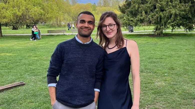 A man and woman smile at the camera in a park. They're both well-dressed.